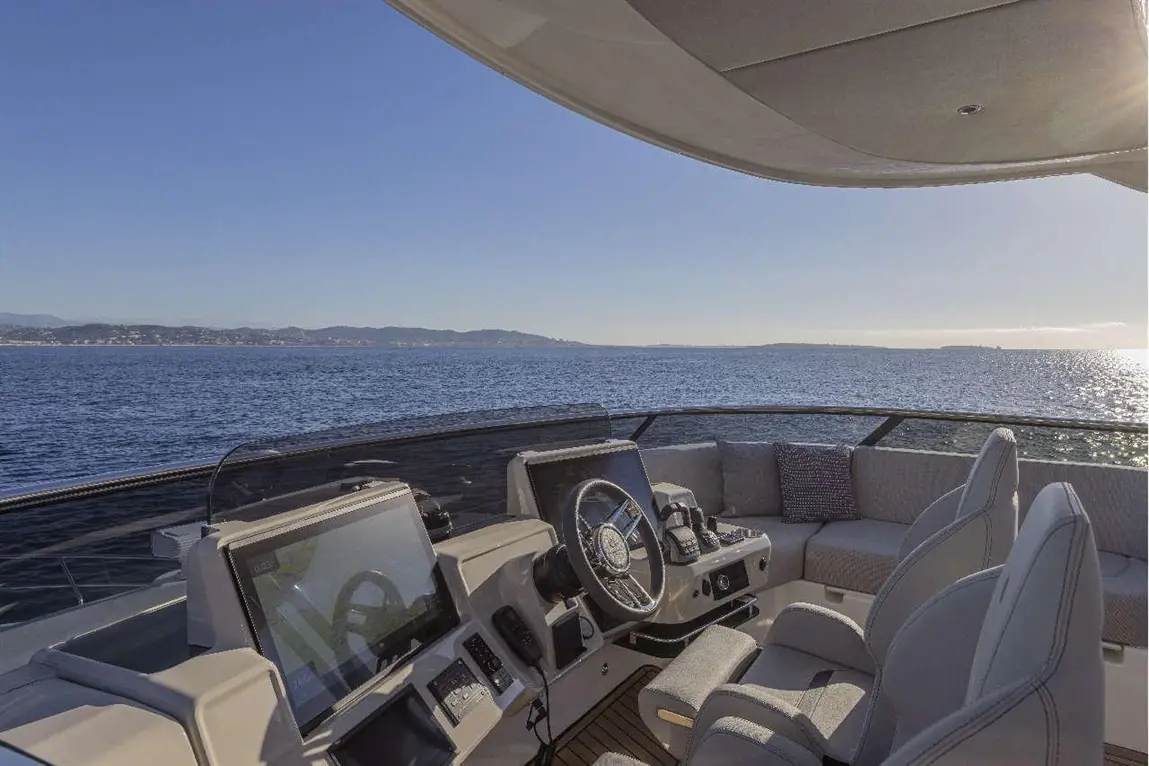 Luxury yacht interior with modern steering wheel, electronic displays, and plush seating overlooking serene blue waters.